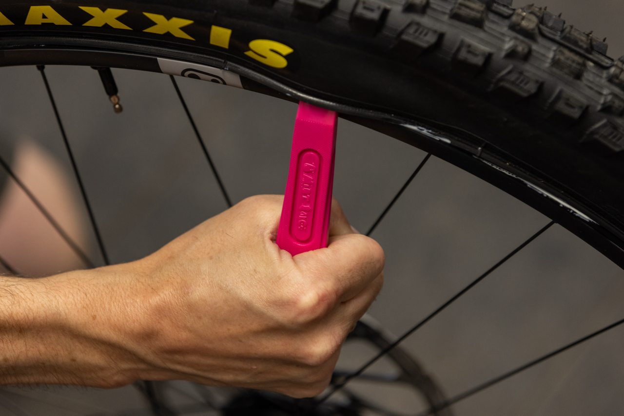 Hand holding a pink tyre lever with Mint Sauce branding near a Maxxis bicycle tyre.
