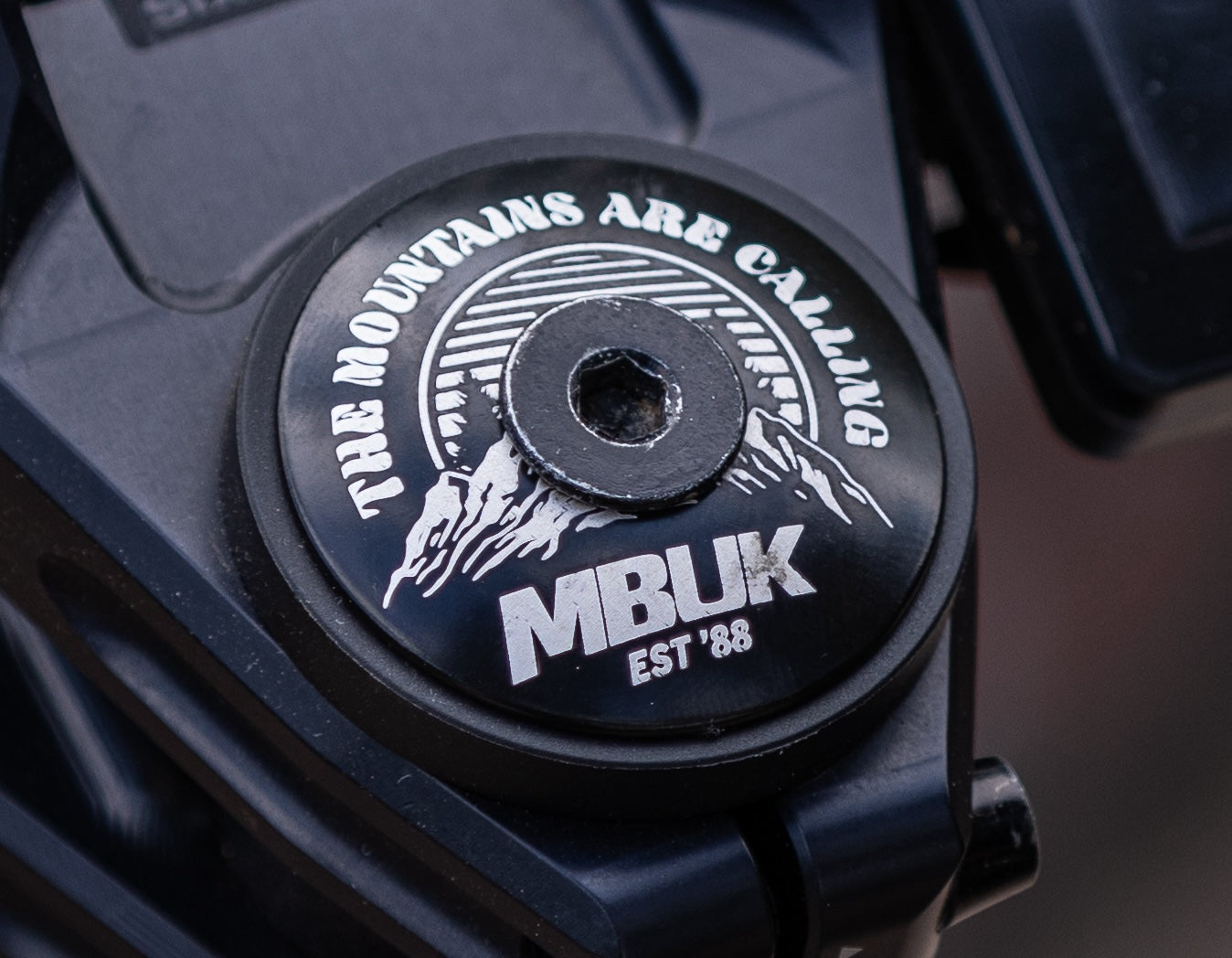 Black stem cap with 'The Mountains Are Calling' and MBUK branding on a dark background